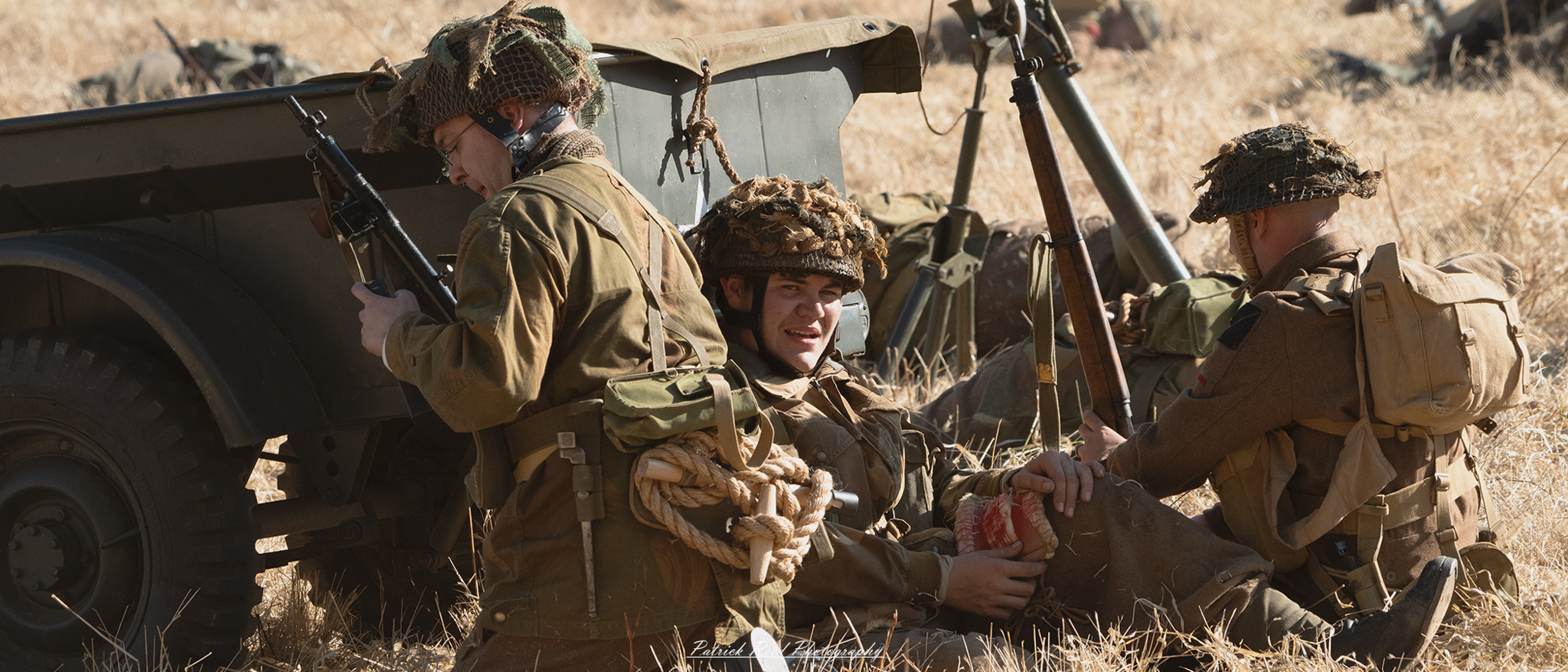 A wounded American soldier leans against a World War II-era jeep, his face displaying exhaustion and pain. His uniform is soiled and torn, with visible bloodstains hinting at the battle he has endured. The jeep, parked on rough terrain, has its canvas top down, exposing its interior filled with gear and supplies. The background captures a war-torn landscape with smoke rising in the distance, emphasizing the chaos of combat. Despite his injuries, the soldier maintains a determined expression, showcasing resilience amidst the harsh realities of war.